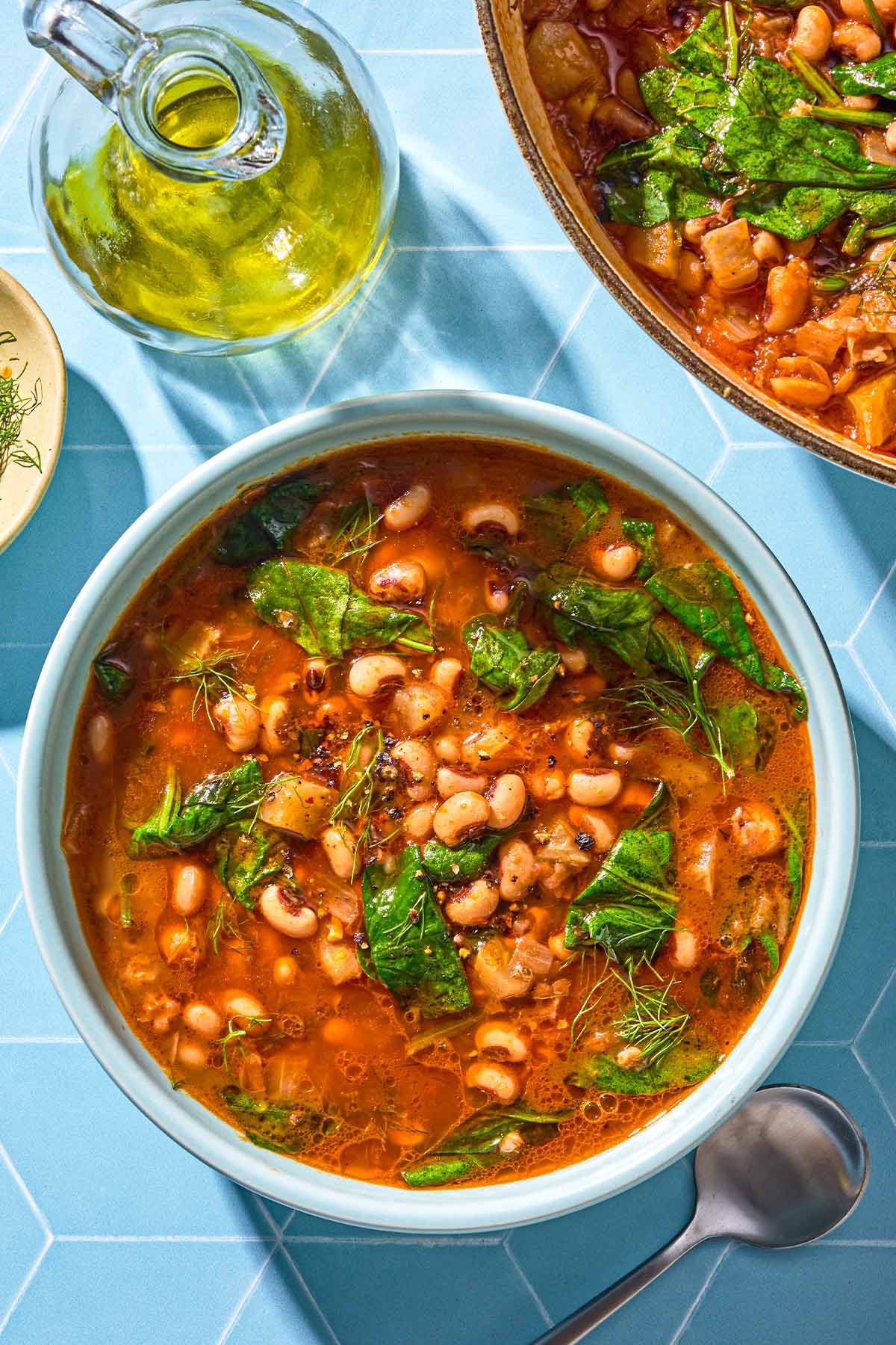 A serving of black eyed peas in a bowl next to a spoon, a bottle of olive oil and the rest of the stew in a pot.