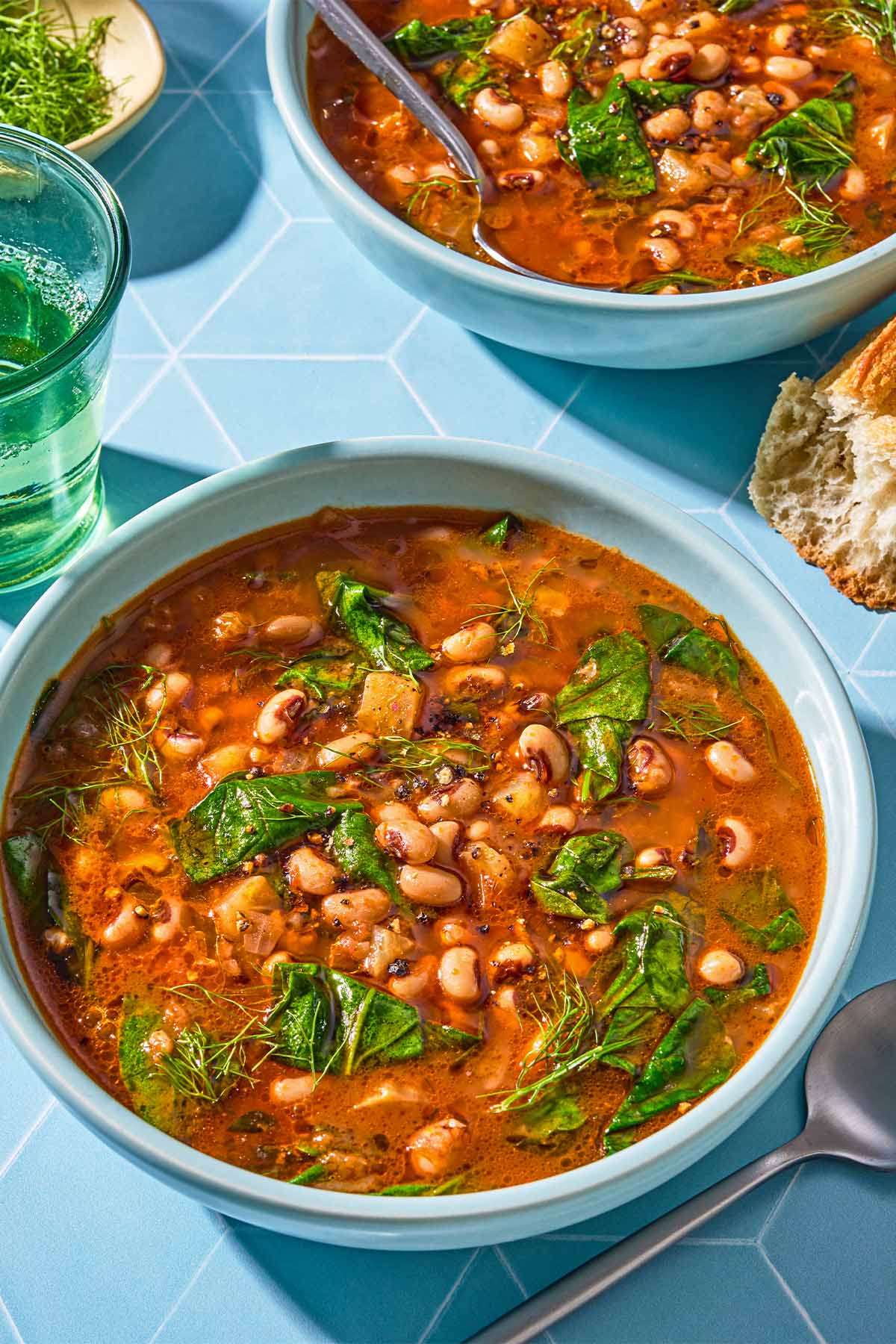 Two servings of black eyed peas in bowls, one with a spoon. Next to these is another spoon, a piece of baguette, a glass of white wine and a bowl of fennel fronds.