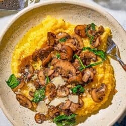 A plate of mushroom polenta with a fork. Next to this is a block of parmesan cheese on a cheese grater, a bowl of black pepper and a bottle of olive oil.