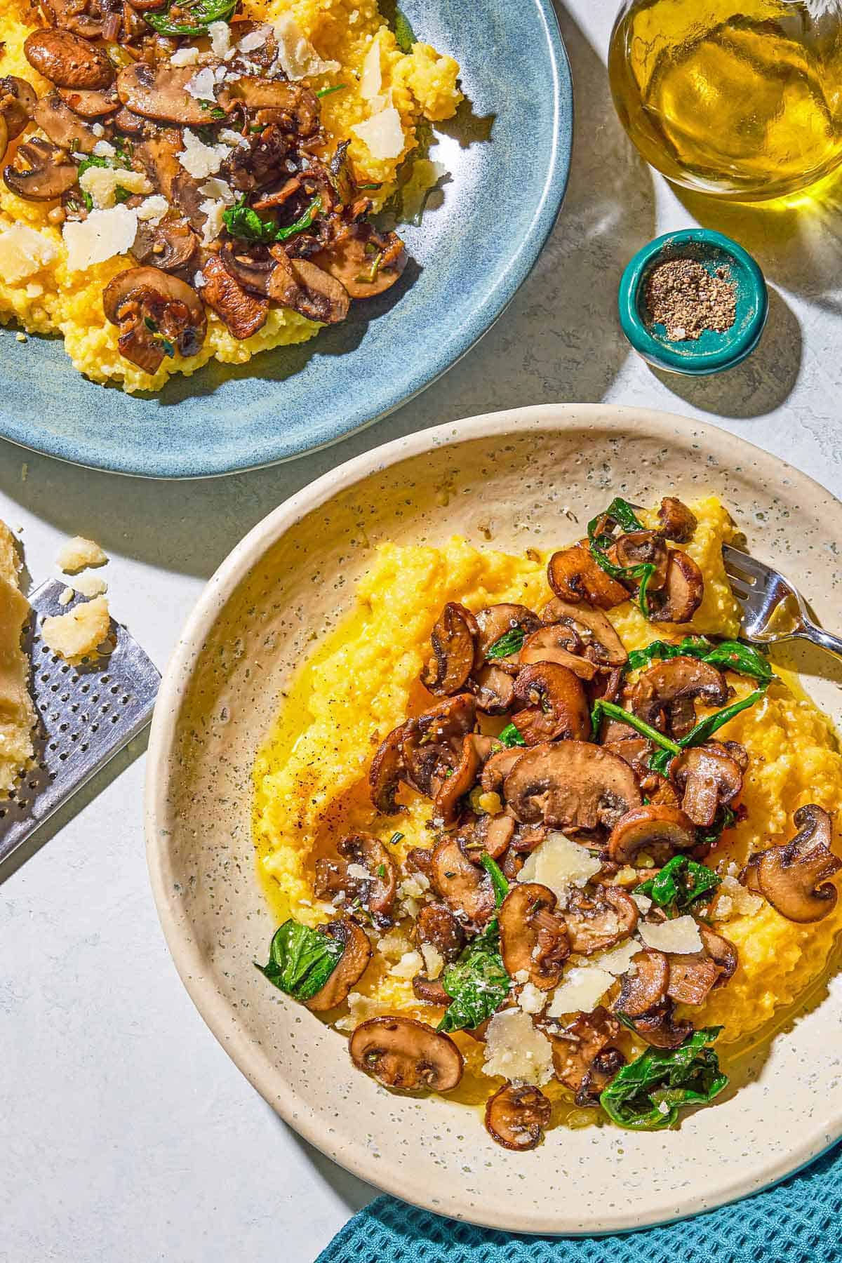 Two plates of mushroom polenta next to a bowl of pepper and a bottle of olive oil.