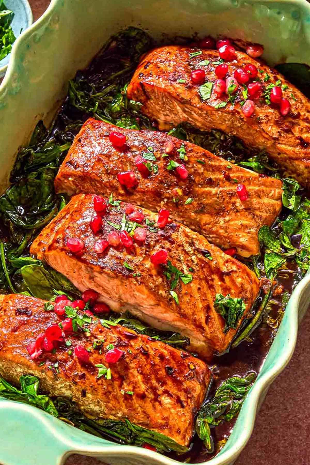 Glazed salmon fillets topped with pomegranate arils on a bed of baby spinach in a baking dish.