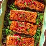 Glazed salmon fillets topped with pomegranate arils on a bed of baby spinach in a baking dish next to a bowl of pomegranate arils.