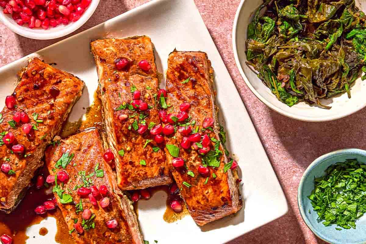 Glazed salmon fillets topped with pomegranate arils on a platter next to a bowls of cooked spinach, more pomegranate arils and herbs.