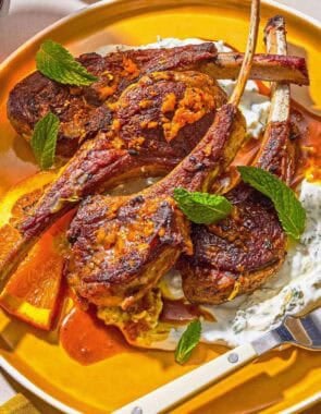 A plate with lamb lollipops, orange wedges, mint yogurt sauce and a fork.