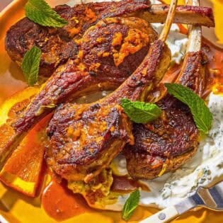 A plate with lamb lollipops, orange wedges, mint yogurt sauce and a fork.