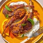 A plate with lamb lollipops, orange wedges, mint yogurt sauce and a fork.
