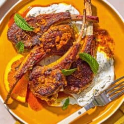 A plate with lamb lollipops, orange wedges, mint yogurt sauce and a fork.