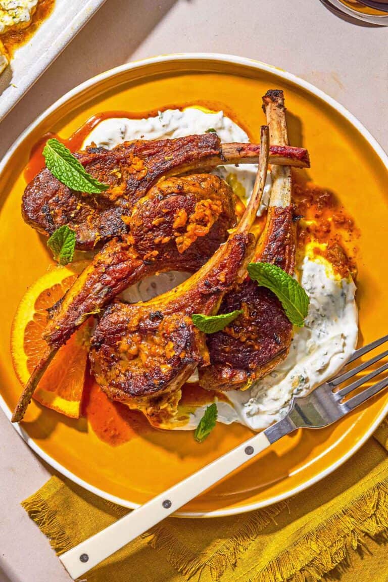 A plate with lamb lollipops, orange wedges, mint yogurt sauce and a fork.