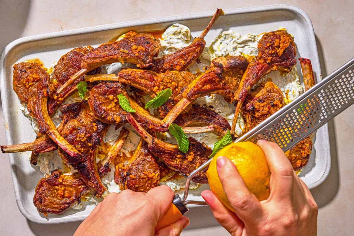 A lemon being zested over a platter of lollipop lamb chops and mint yogurt sauce.