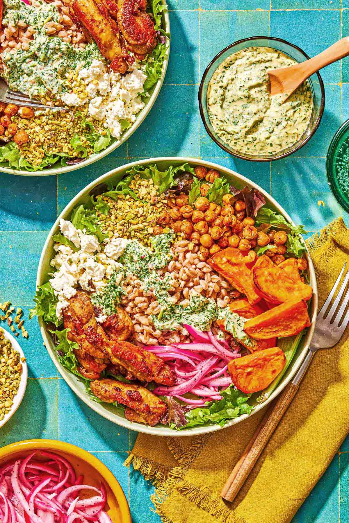 A sweet potato bowl next to a fork on a napkin. Next to this is another sweet potato bowl with a fork, and bowls of green goddess tahini dressing, chopped pistachios and pickled red onions.