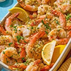 Shrimp oreganata with lemon wedges in a baking dish next to bowls of chopped parsley and red pepper flakes.