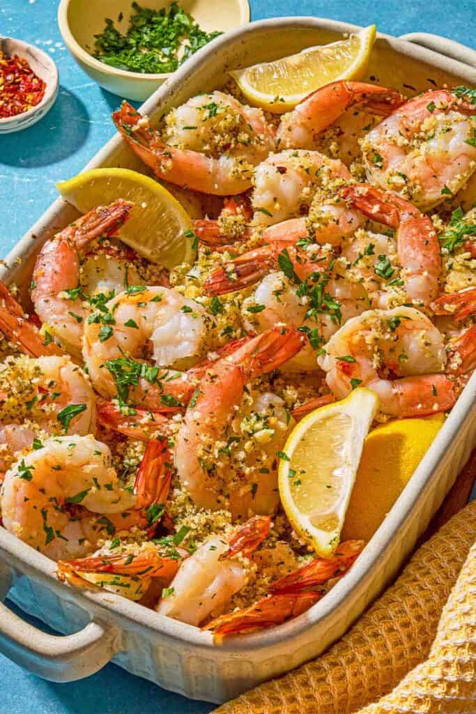 Shrimp oreganata with lemon wedges in a baking dish next to bowls of chopped parsley and red pepper flakes.