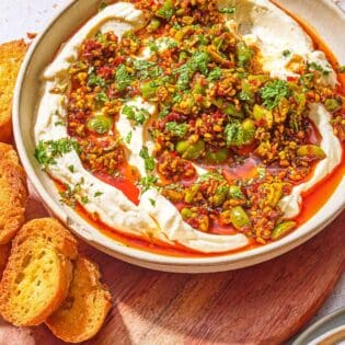Whipped ricotta dip with pistachios, olives and harissa oil in a bowl on a wooden serving board with slices of toasted bread.