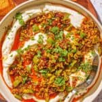 Whipped ricotta dip with pistachios, olives and harissa oil in a bowl on a wooden serving board with slices of toasted bread.