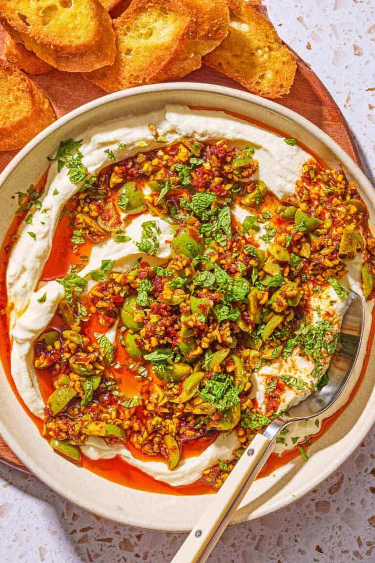 Whipped ricotta dip with pistachios, olives and harissa oil in a bowl on a wooden serving board with slices of toasted bread.