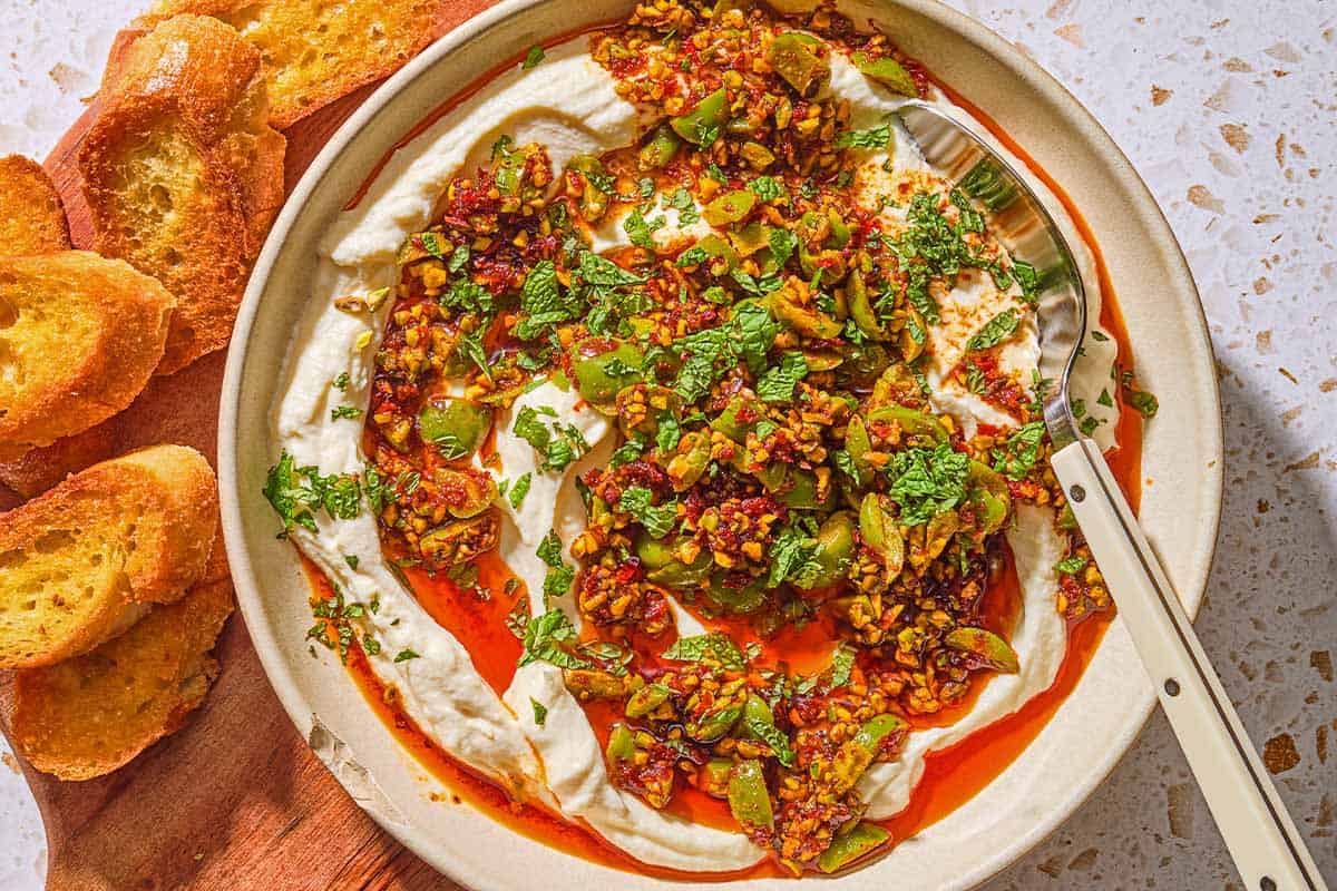 Whipped ricotta dip with pistachios, olives and harissa oil in a bowl with a spoon on a wooden serving board with slices of toasted bread.