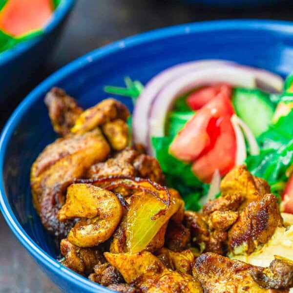 A close up of a chicken shawarma bowl with shawarma chicken, onion, tomatoes, and arugula in a blue bowl against a dark background.