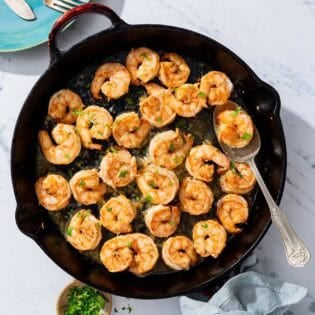 Honey garlic shrimp in a cast iron skillet with a spoon. Next to this is a plate with 2 forks and two knives, a small bowl of chopped parsley and cilantro, and a cloth napkin.