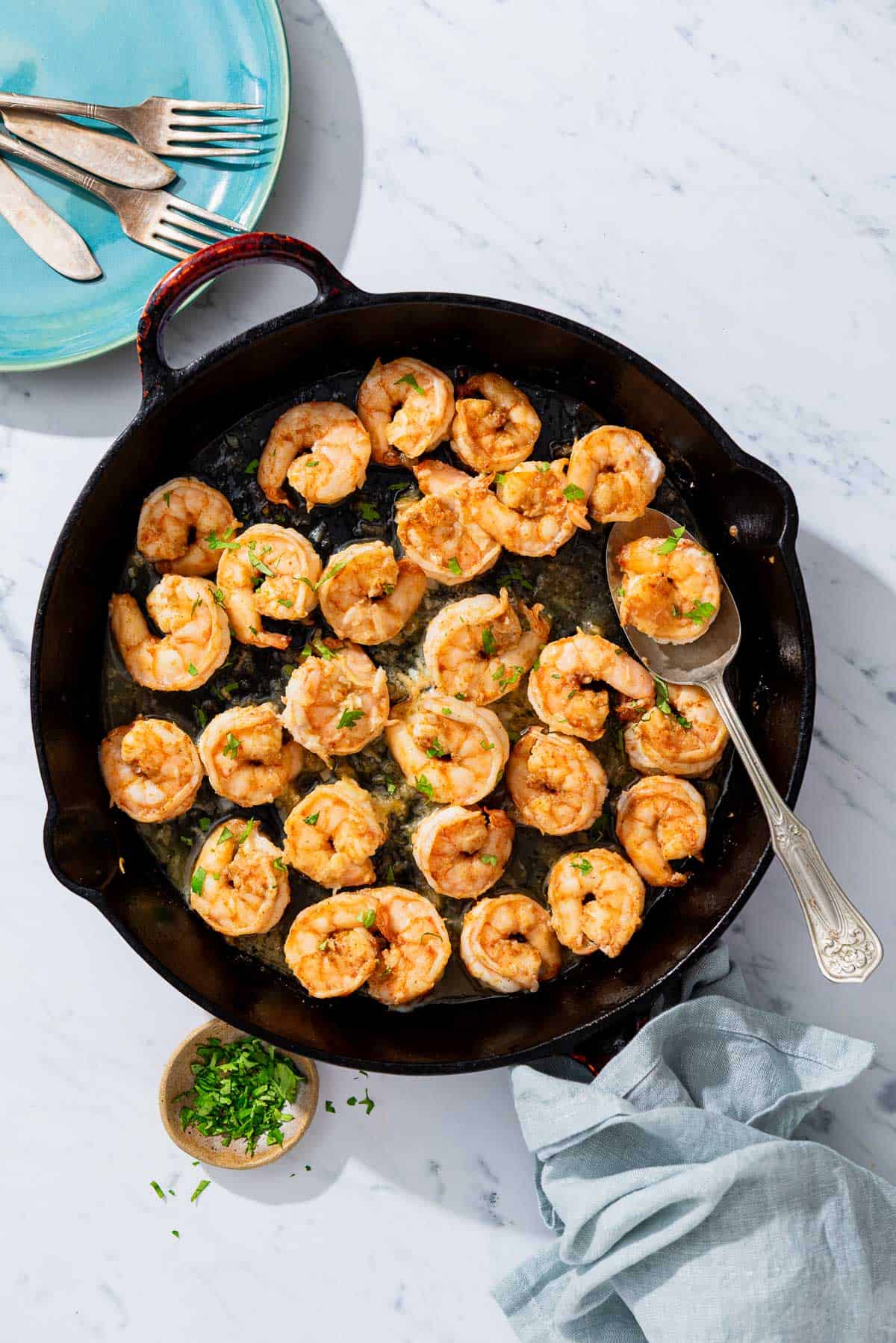 Honey garlic shrimp in a cast iron skillet with a spoon. Next to this is a plate with 2 forks and two knives, a small bowl of chopped parsley and cilantro, and a cloth napkin.
