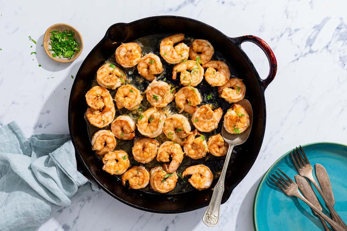 Honey garlic shrimp in a cast iron skillet with a spoon. Next to this is a plate with 2 forks and two knives, a small bowl of chopped parsley and cilantro, and a cloth napkin.