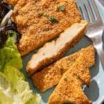A partially sliced parmesan crusted chicken fillet on a plate with a salad and a fork.