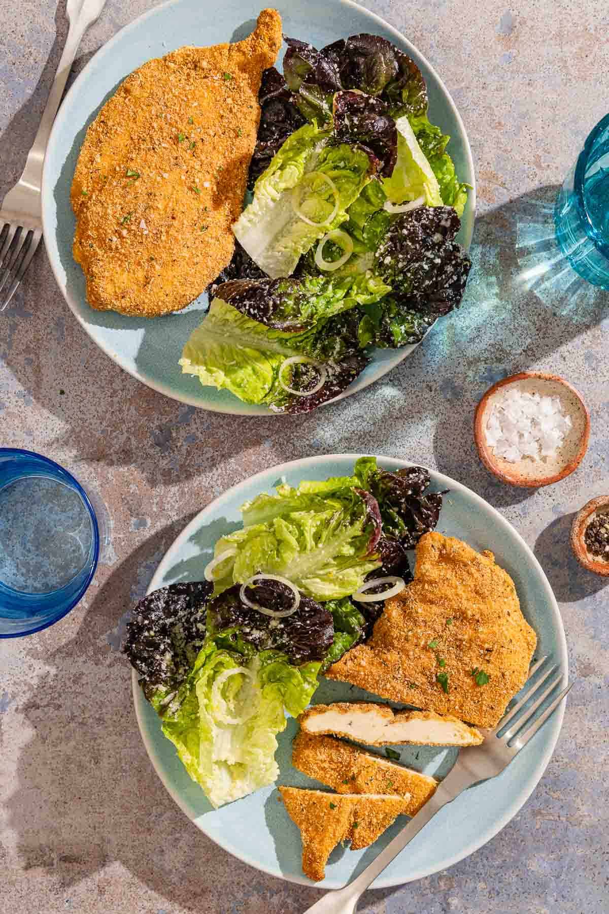 Two plates, each with a parmesan crusted chicken fillet and salad, one with a fork. Next to these are glasses of water, bowls of salt and pepper and another fork.
