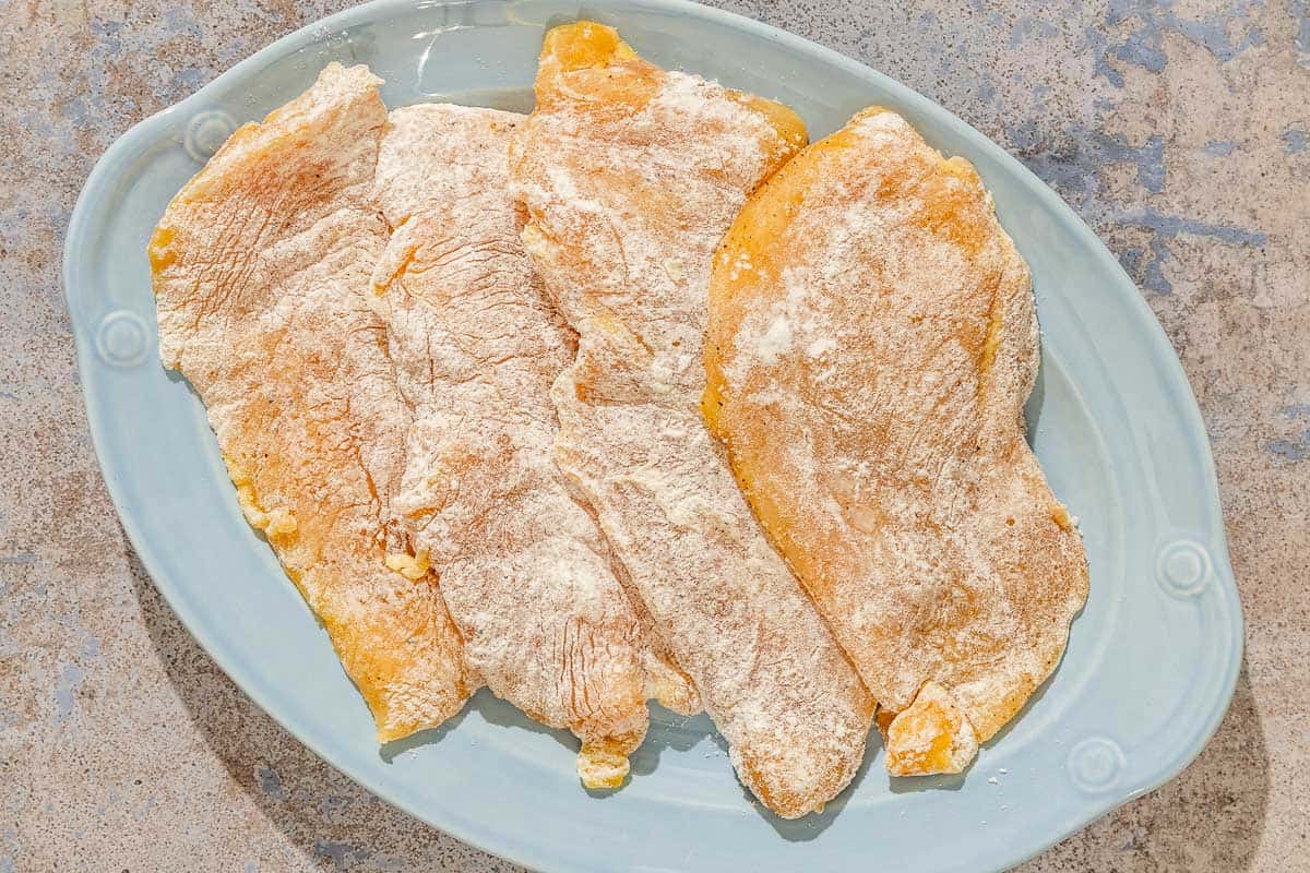 Four uncooked, floured chicken fillets on a platter.