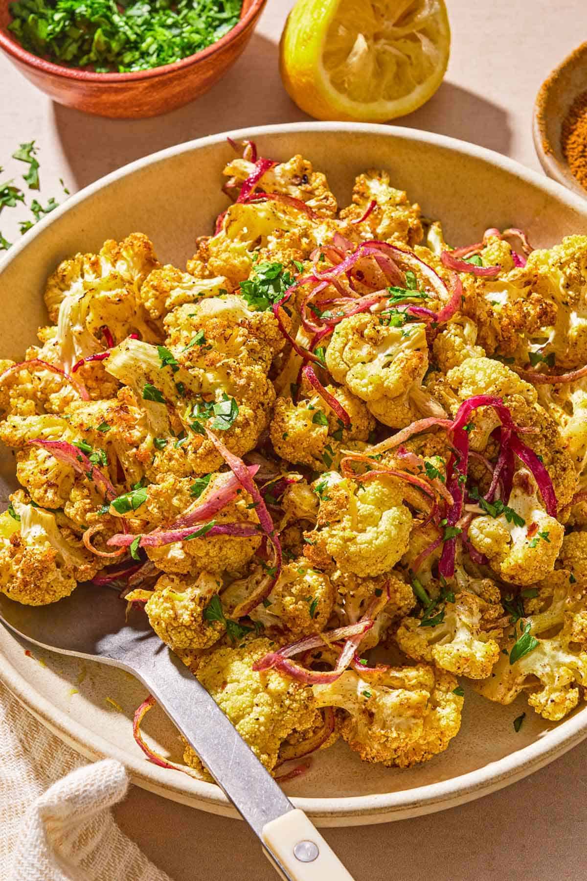 Air fryer cauliflower in a serving bowl with a serving spoon next to bowls of spices and chopped cilantro and a juiced lemon half.