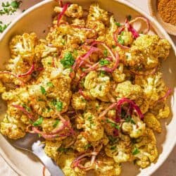 Air fryer cauliflower in a serving bowl with a serving spoon next to bowls of spices and chopped cilantro and a juiced lemon half.