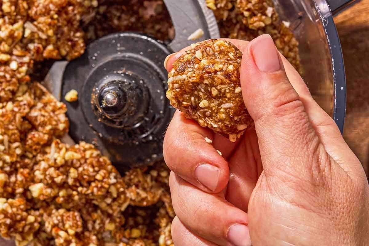 An energy ball being formed over the energy ball batter in a the bowl of a food processor fitted with a blade.