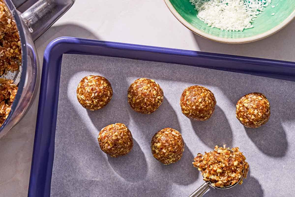 Six energy balls with a scoop of the batter on a parchment lined sheet pan. Next to this is a bowl of shredded coconut and the rest of the batter in the bowl of a food processor.