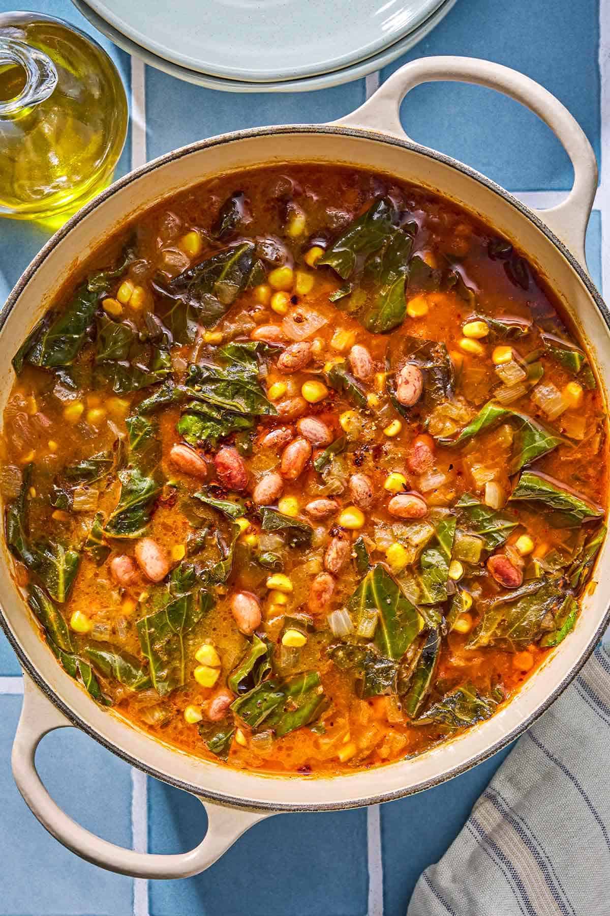Collard green soup in a pot next to a bottle of olive oil, a kitchen towel and a stack of 2 bowls.