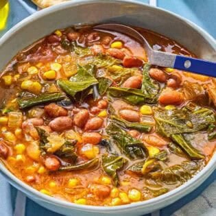 Collard green soup in a bowl with a spoon. Next to this is a kitchen towel and pieces of pita bread.