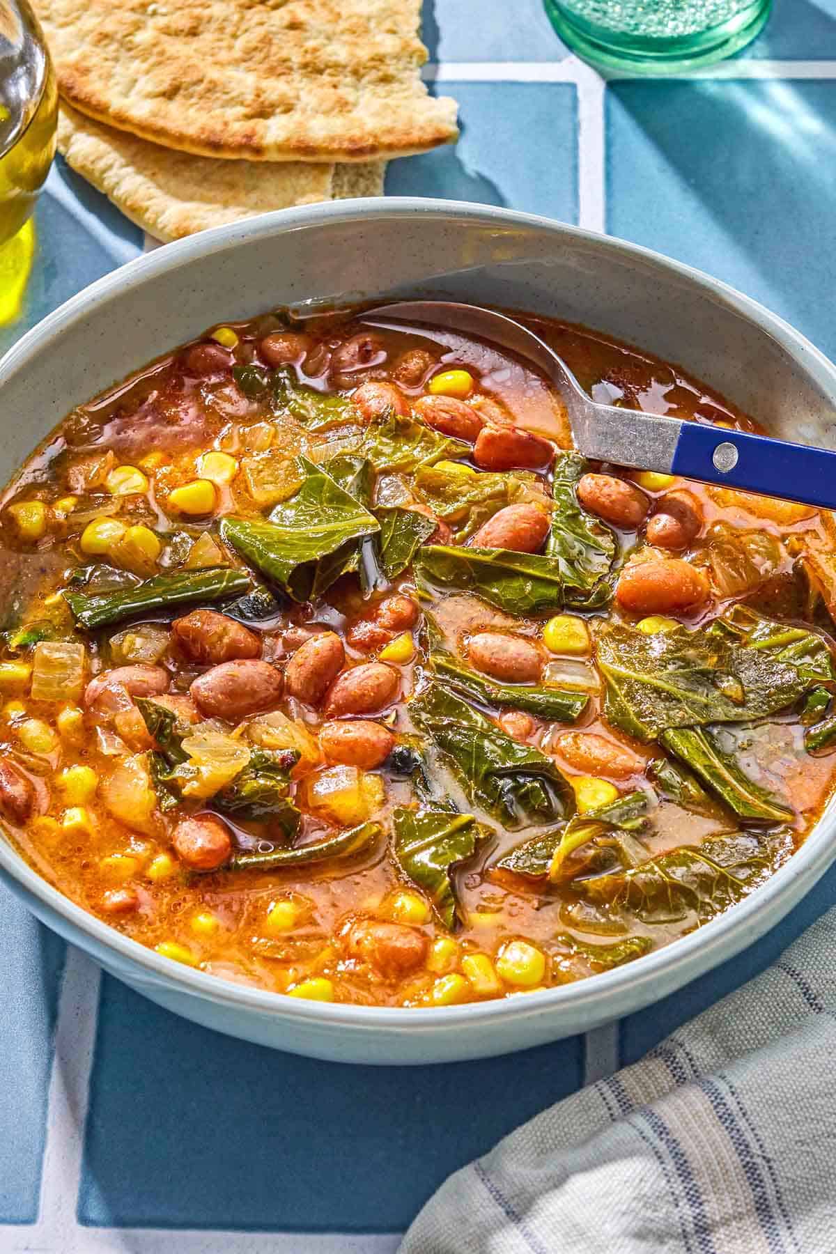 Collard green soup in a bowl with a spoon. Next to this is a kitchen towel and pieces of pita bread.