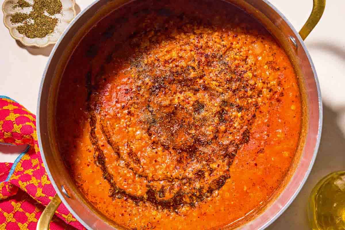 Turkish lentil stew topped with infused olive oil in a large pot next to a bowl of spices and a cloth napkin.