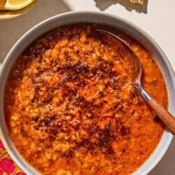 Turkish lentil stew topped with infused olive oil in a bowl with a spoon next to pieces of flatbread and a bowl of lemon wedges.