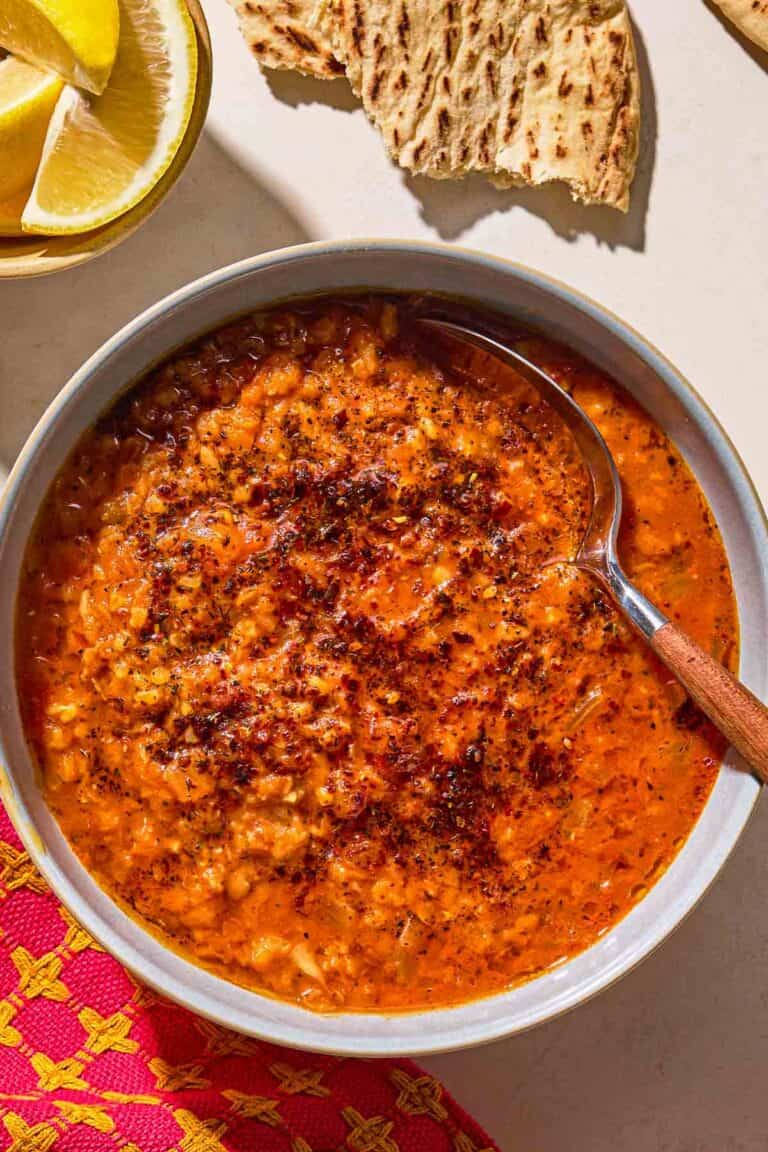 Turkish lentil stew topped with infused olive oil in a bowl with a spoon next to pieces of flatbread and a bowl of lemon wedges.