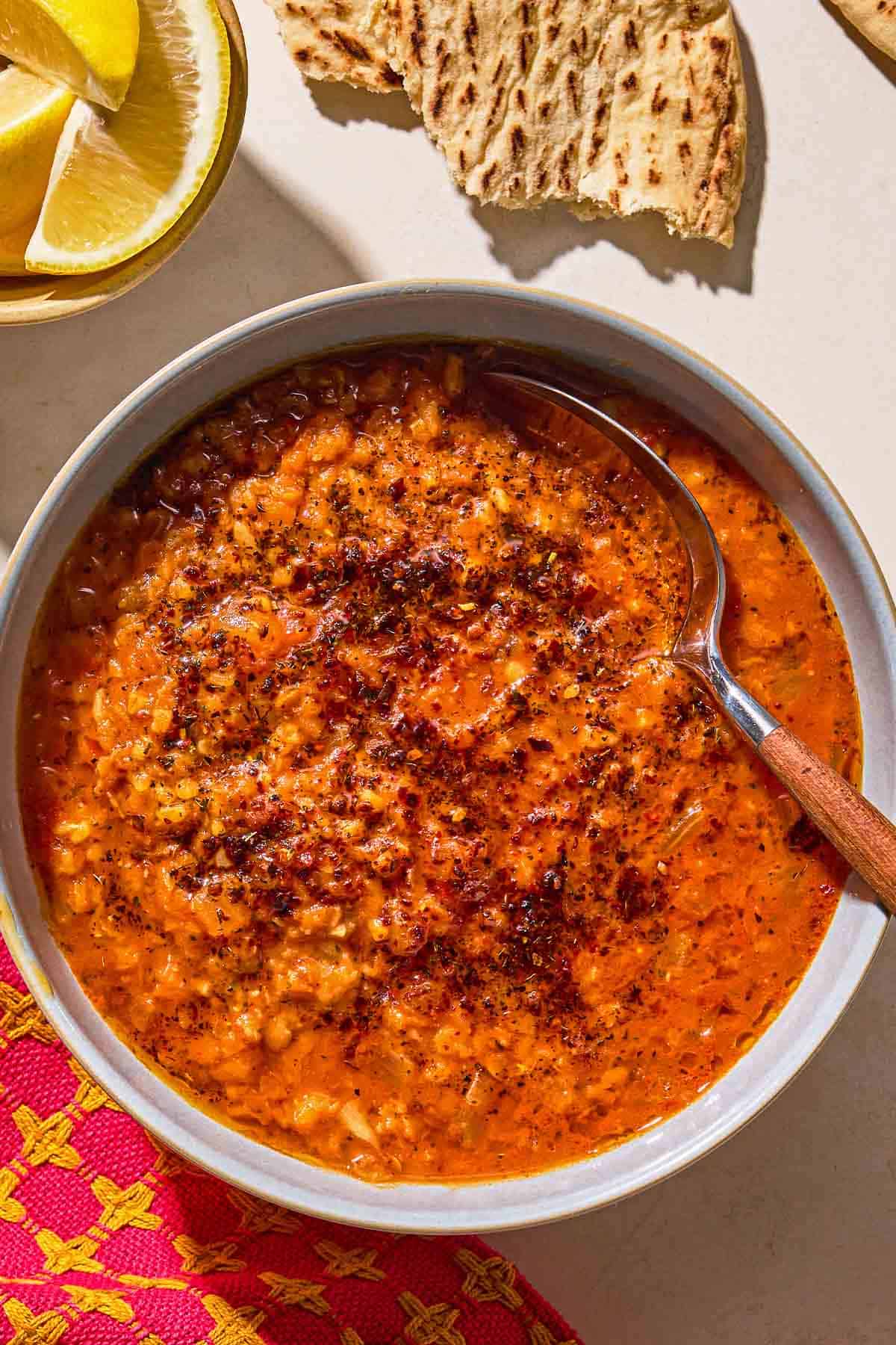 Turkish lentil stew topped with infused olive oil in a bowl with a spoon next to pieces of flatbread and a bowl of lemon wedges.