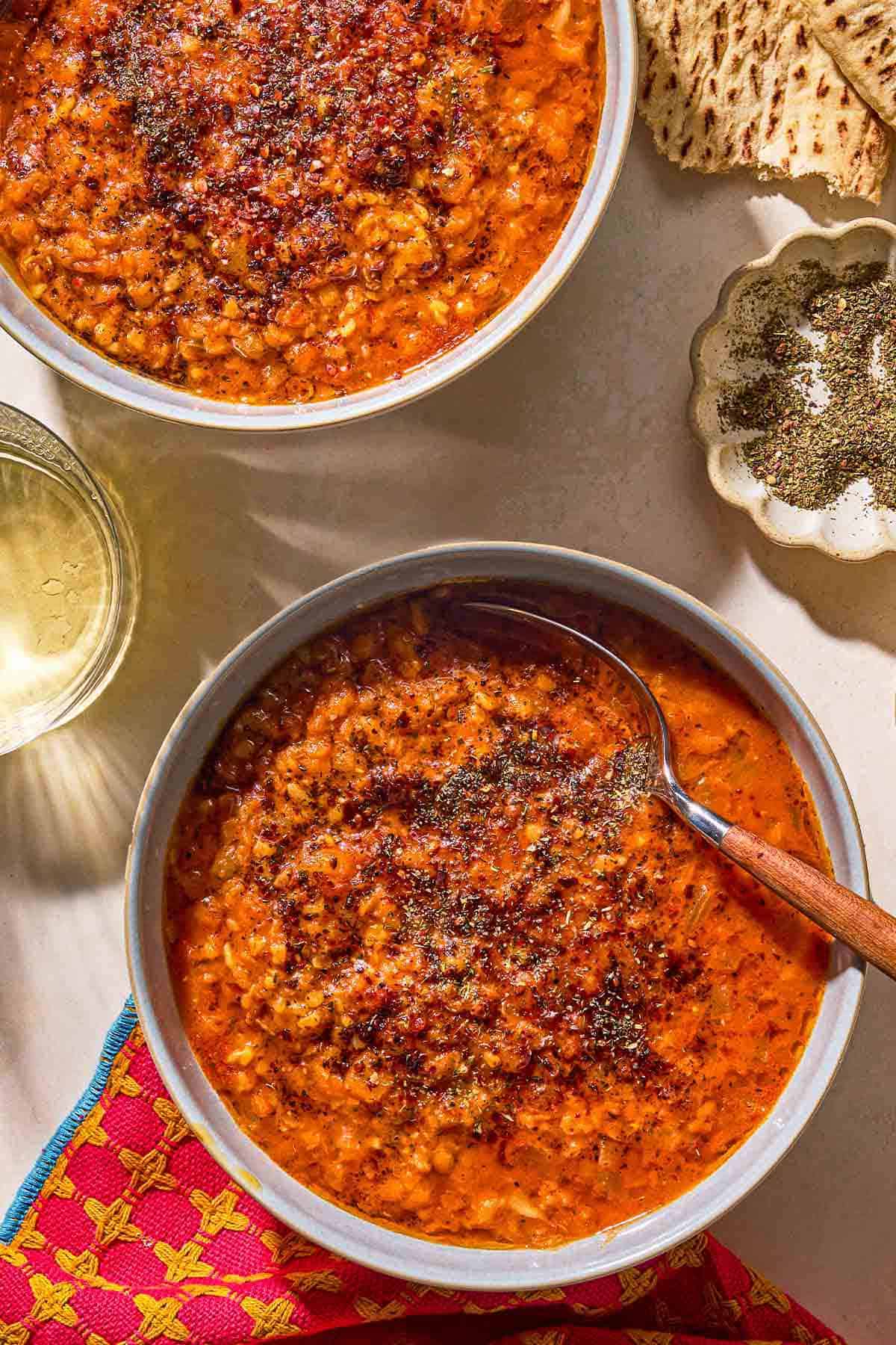 Turkish lentil stew topped with infused olive oil in a bowl with a spoon. Next to this is another bowl of the stew, a cloth napkin, a bowl of spices, pieces of flatbread, and a glass of wine.