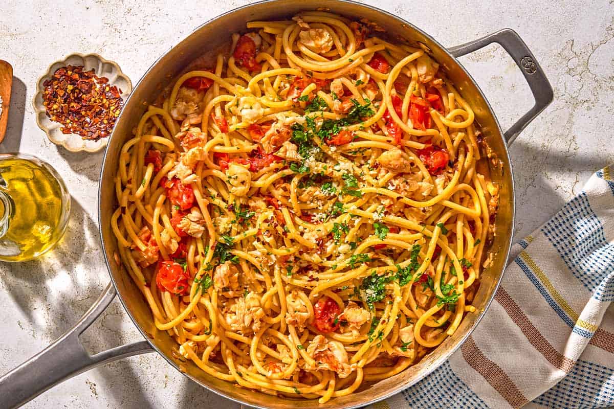 Lobster pasta in a tall sided skillet next to a bowl of red pepper flakes, a bottle of olive oil and a kitchen towel.