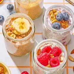 Four jars of overnight oats with various toppings. Next to these are raspberries, blueberries, almonds and a spoon.