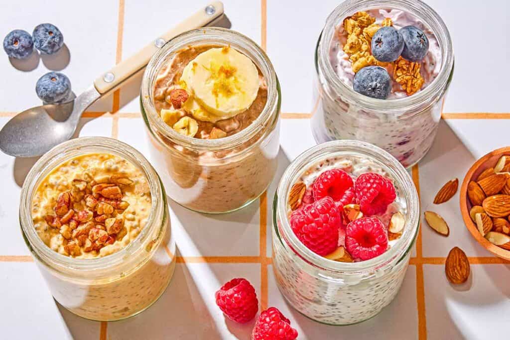 Four jars of overnight oats with various toppings. Next to these are raspberries, blueberries, almonds and a spoon.