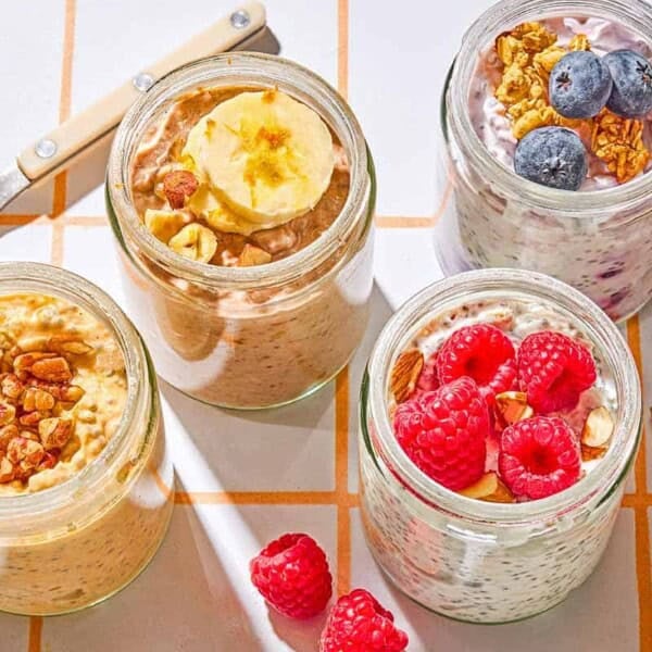 Four jars of overnight oats with various toppings. Next to these are raspberries, blueberries, almonds and a spoon.