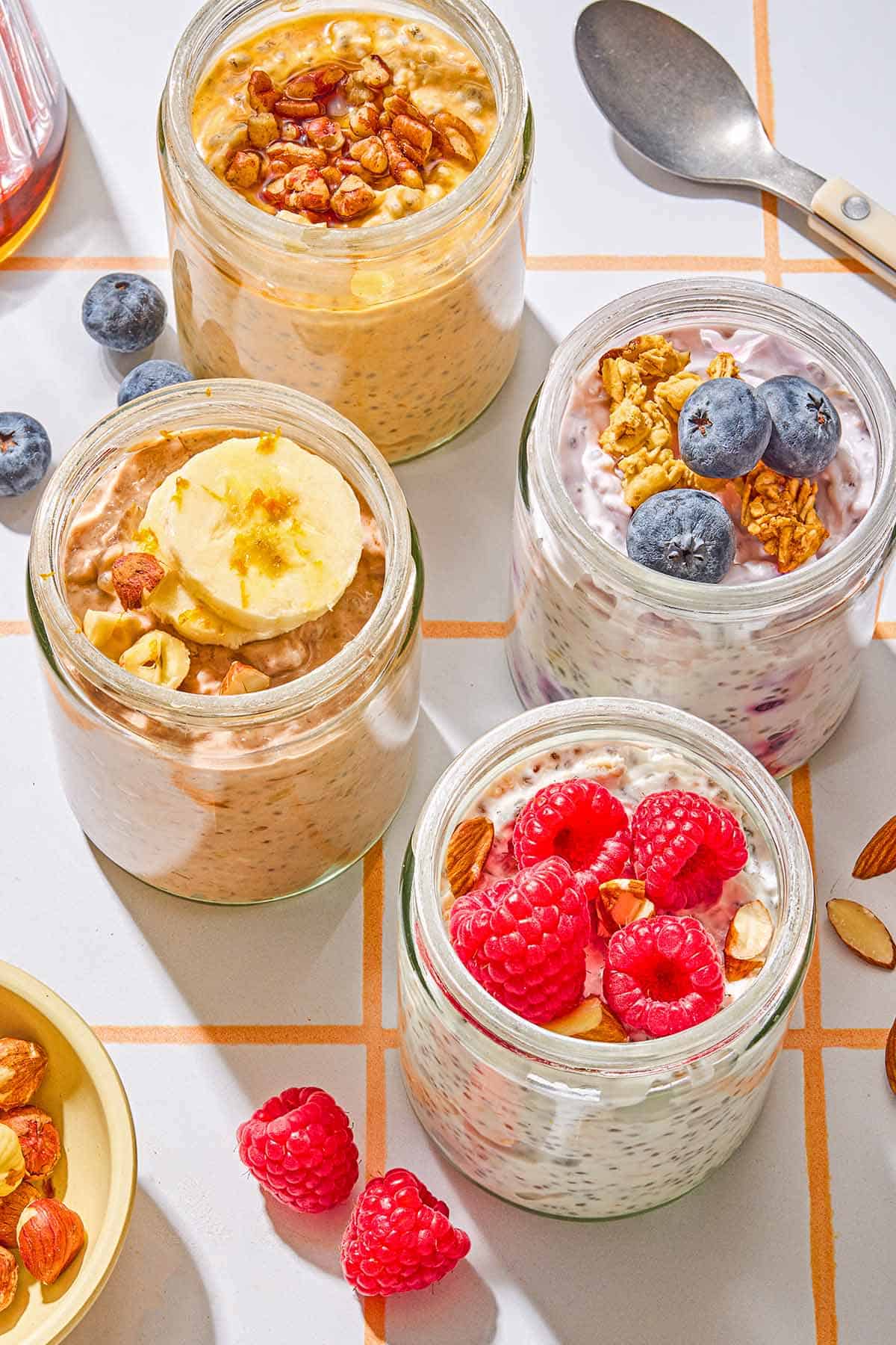 Four jars of overnight oats with various toppings. Next to these are raspberries, blueberries, almonds and a spoon.