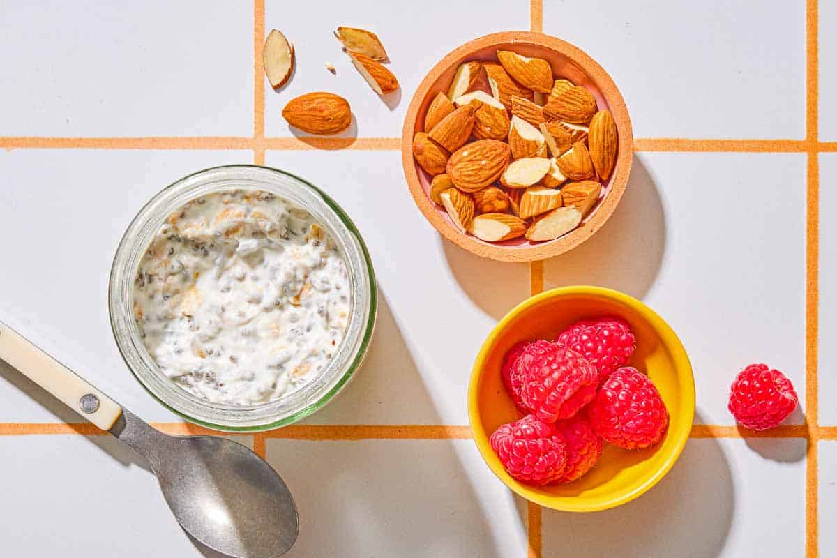 A jar of overnight oats next to a spoon, and bowls of almonds and raspberries.