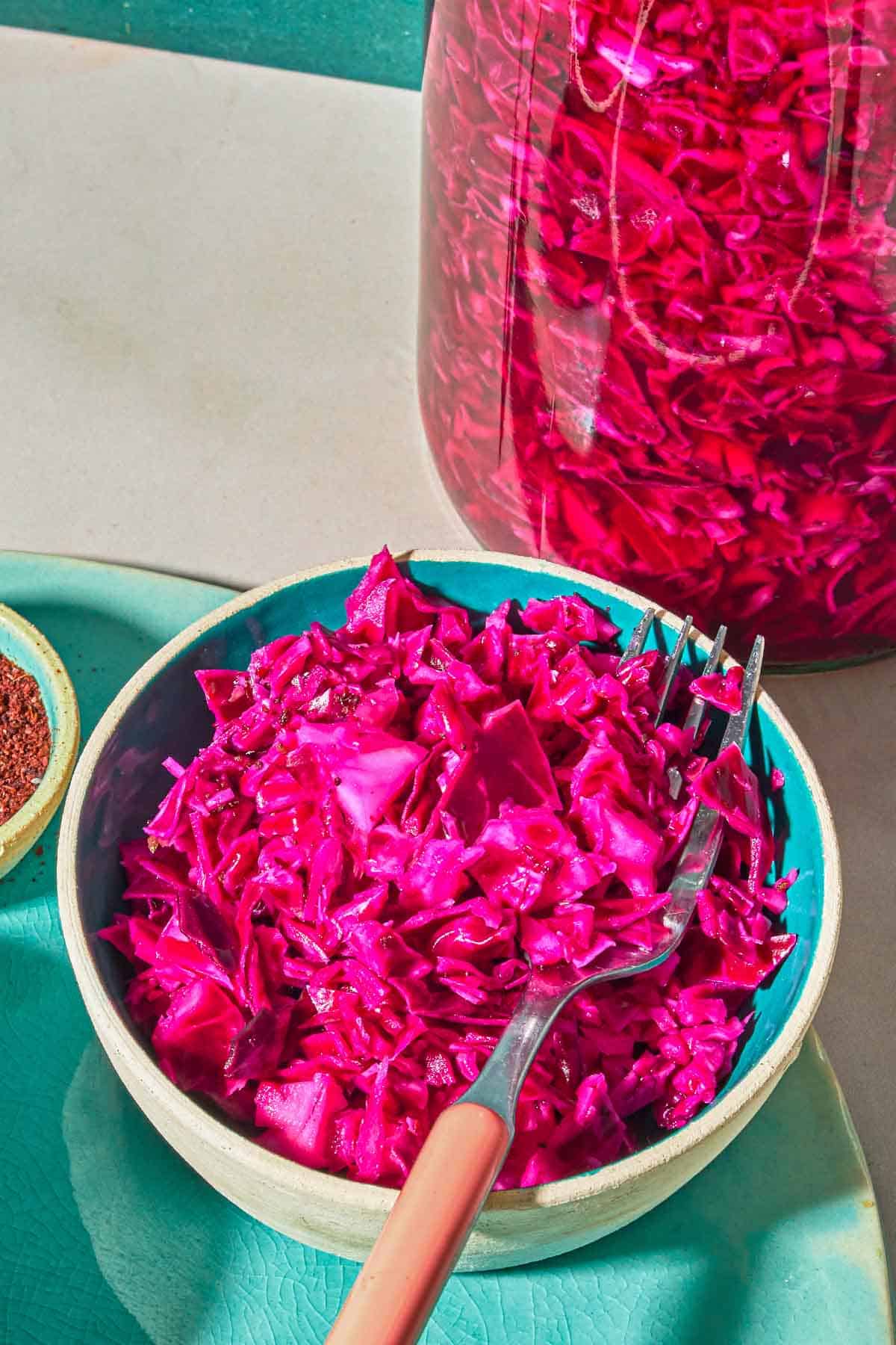 A bowl of pickled cabbage with a fork next to a jar of the pickled cabbage and a bowl of sumac.