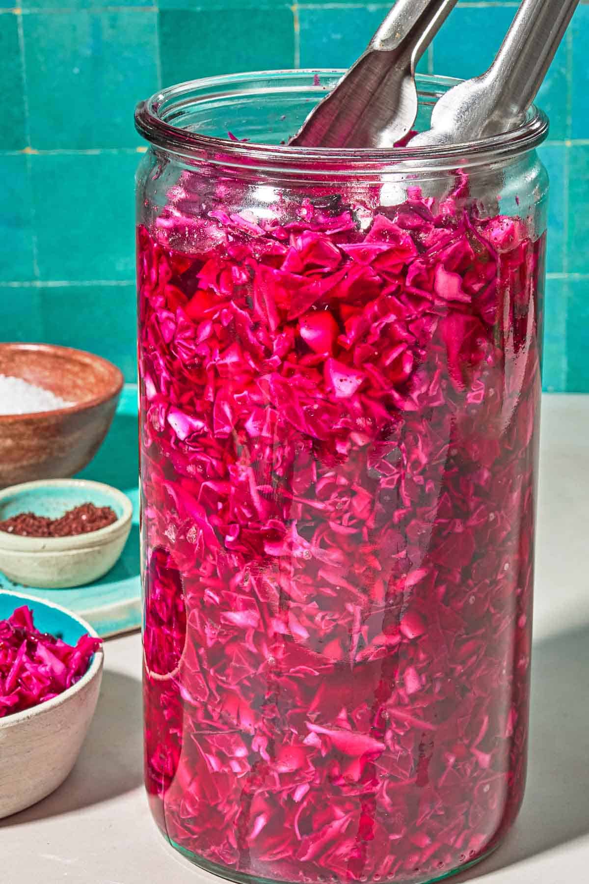 Pickled cabbage in a jar with a pair of tongs next to bowls of pickled cabbage, sumac and salt.