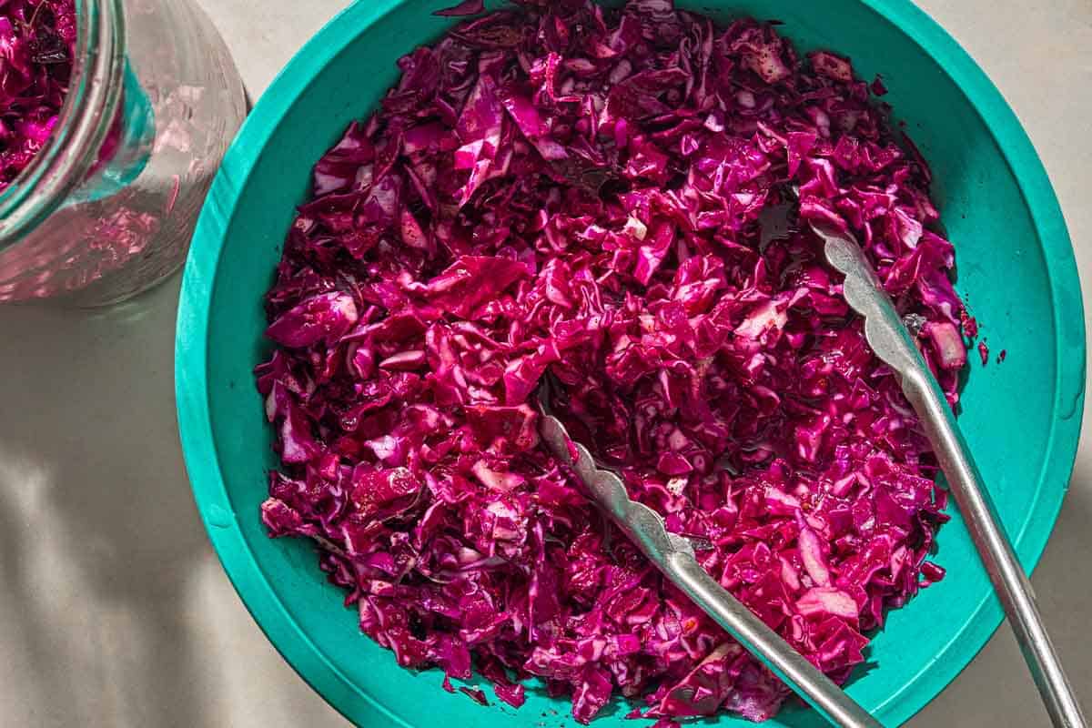 The chopped cabbage for the pickled cabbage in a bowl with a pair of tongs next to a jar with more of the chopped cabbage.