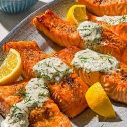 Six lemon dill salmon fillets on a platter with lemon wedges. Next to this is a bowl of dill sauce for salmon with a spoon.