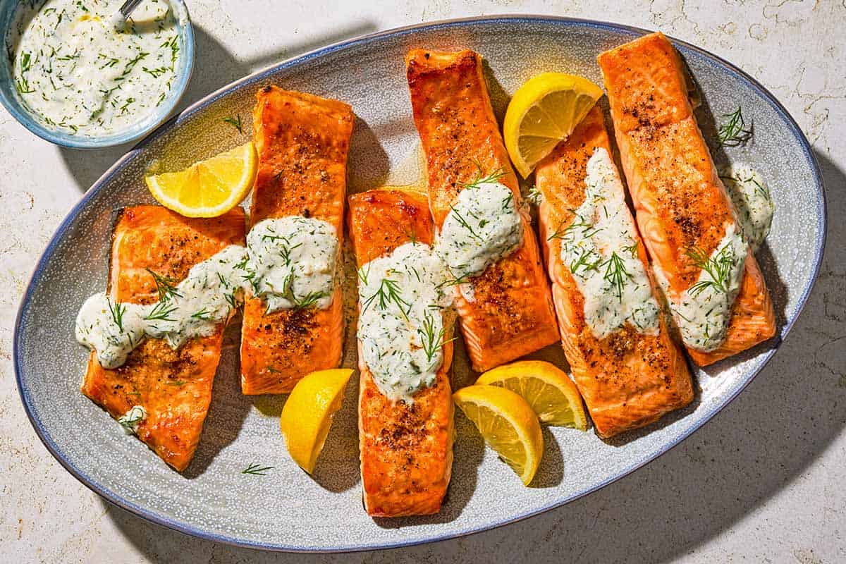 Six lemon dill salmon fillets on a platter with lemon wedges next to a bowl of dill sauce for salmon.